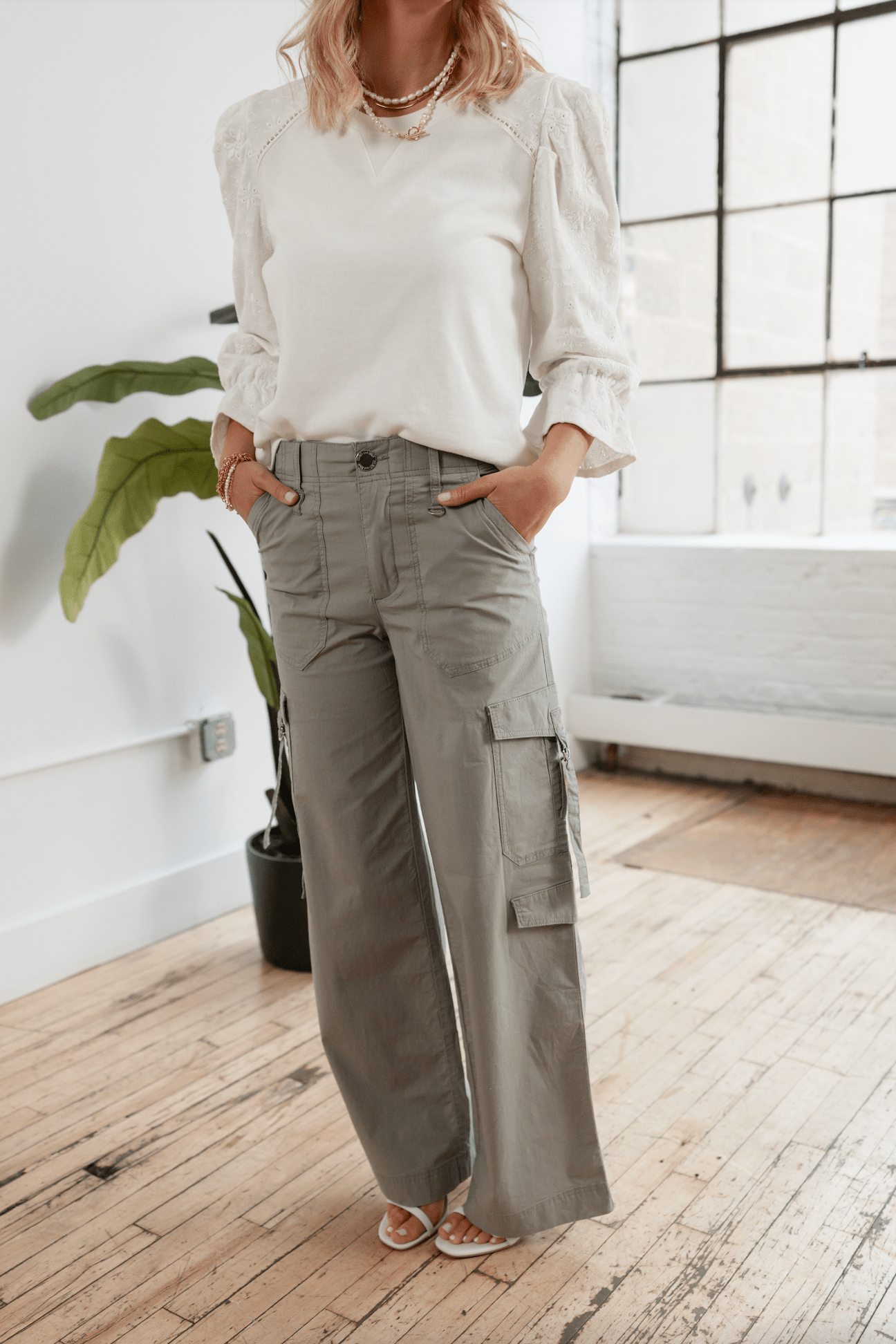 Woman in a white blouse and sage cargo pants standing in a room with a plant and clothing rack.